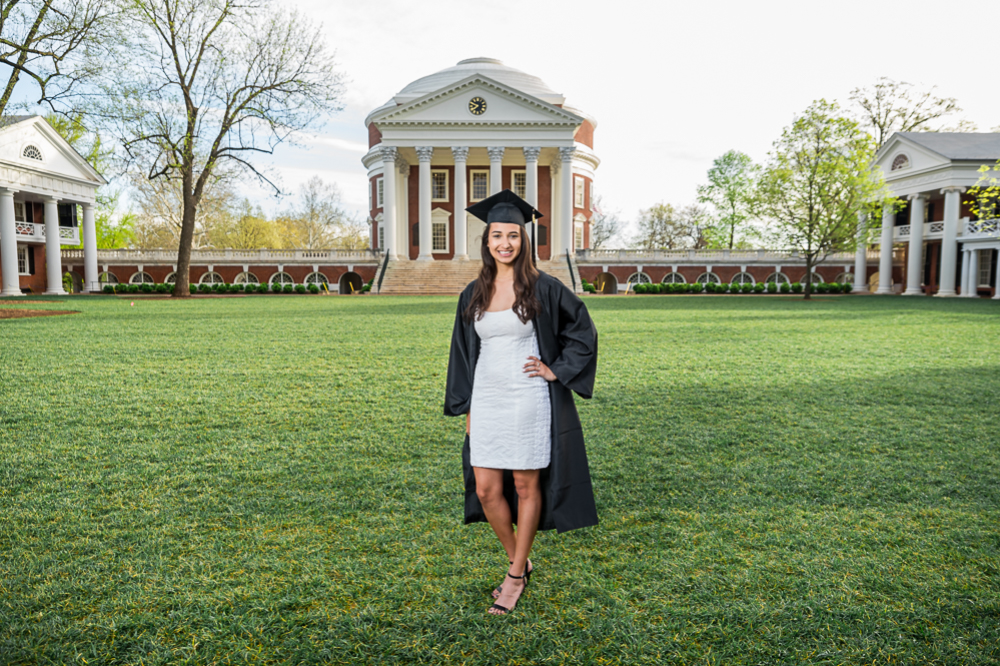 A young woman wearing a cap and gown smiles during her grad photoshoot on UVA's lawn in front of the Rotunda