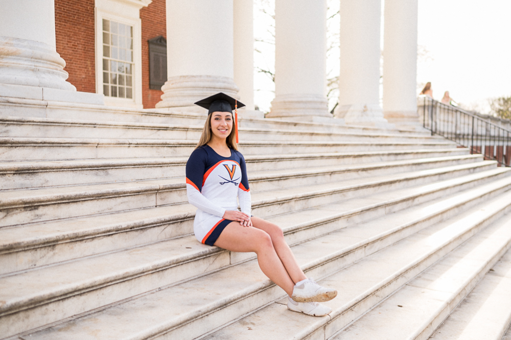A UVA Cheerleader sits on the Rotunda Steps in her cheer uniform and a graduation cap, posing for her grad photos