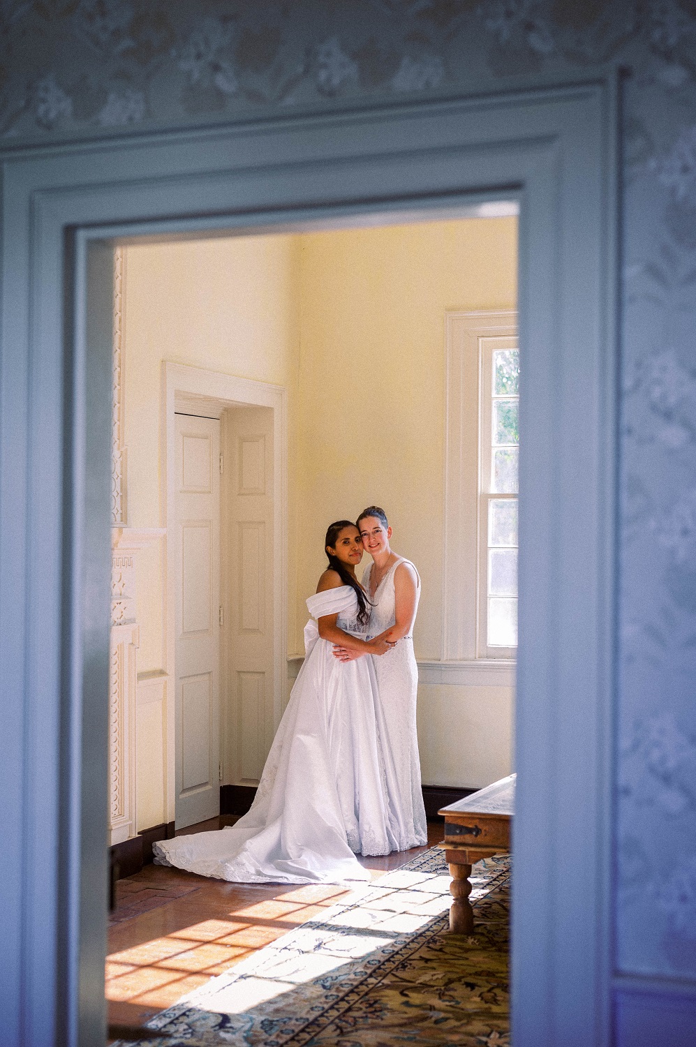 Two women wearing white dresses hold each other on during their same-sex hispanic-american wedding day, inside the parlor of a historic mansion in Maryland, VA. Photographed by Hunter and Sarah Photography.