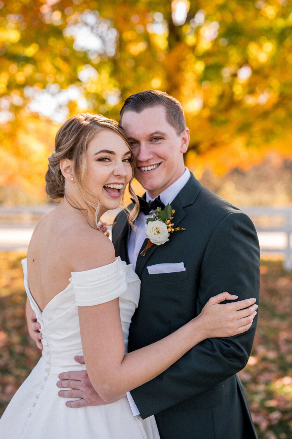 A bride in a white dress and a groom in a green velvet suit smile brightly with vibrant yellow foliage behind them during their wedding day at Castle Hill Cider in Charlottesville, VA. Beautifully captured by Hunter and Sarah Photography.