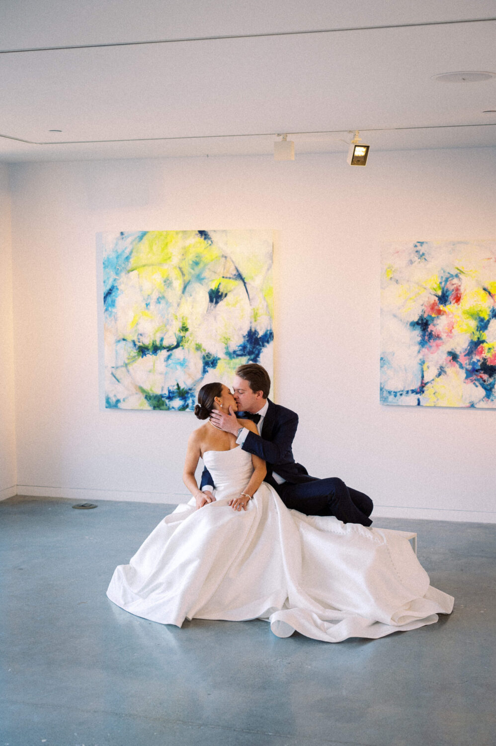 A bride and groom sit inside an art gallery during their Doyle Hotel Wedding, elegantly kissing with art on the walls behind them. Beautifully captured by Hunter and Sarah Photography