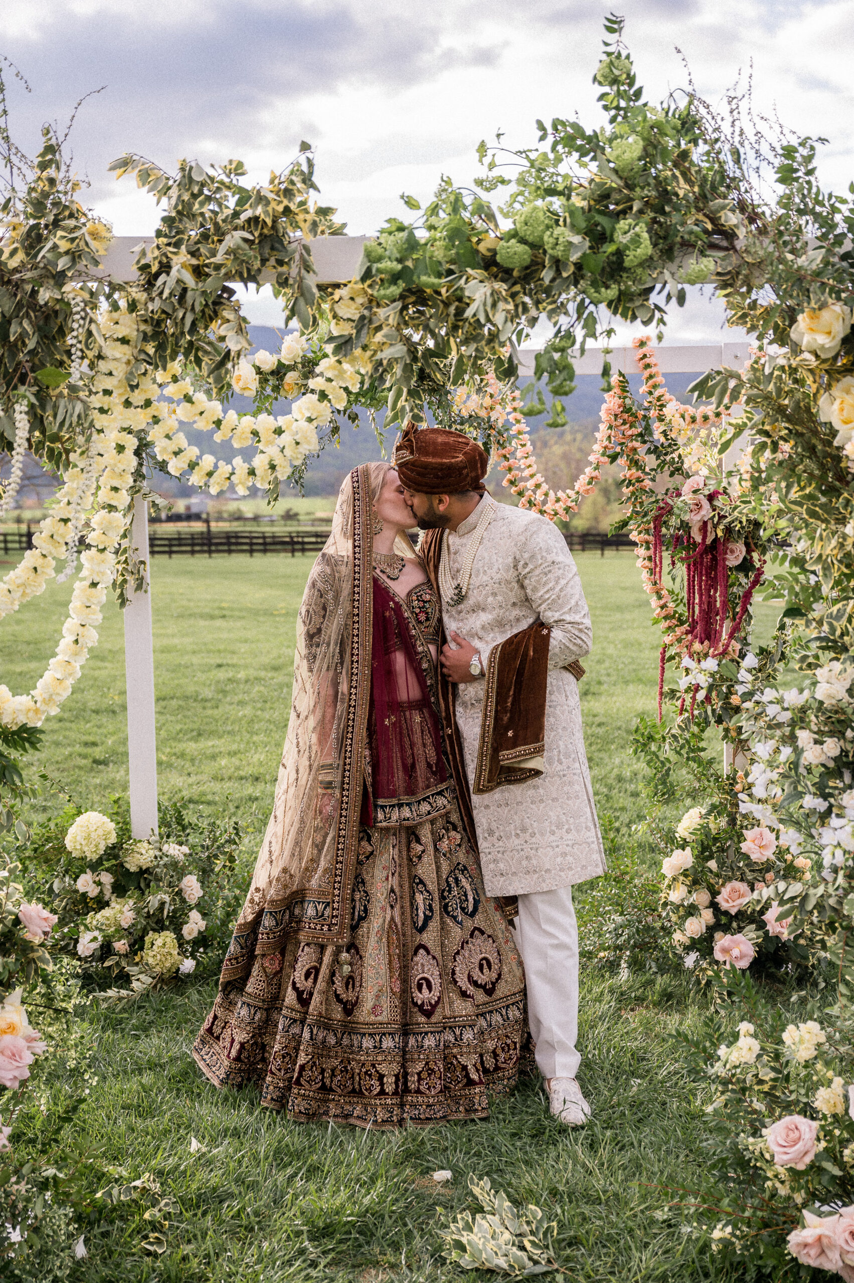A white American bride and an Indian-American groom kiss under a floral-covered mandap, during their Indian fusion wedding at King Family Vineyards, expertly photographed by Hunter and Sarah Photography.
