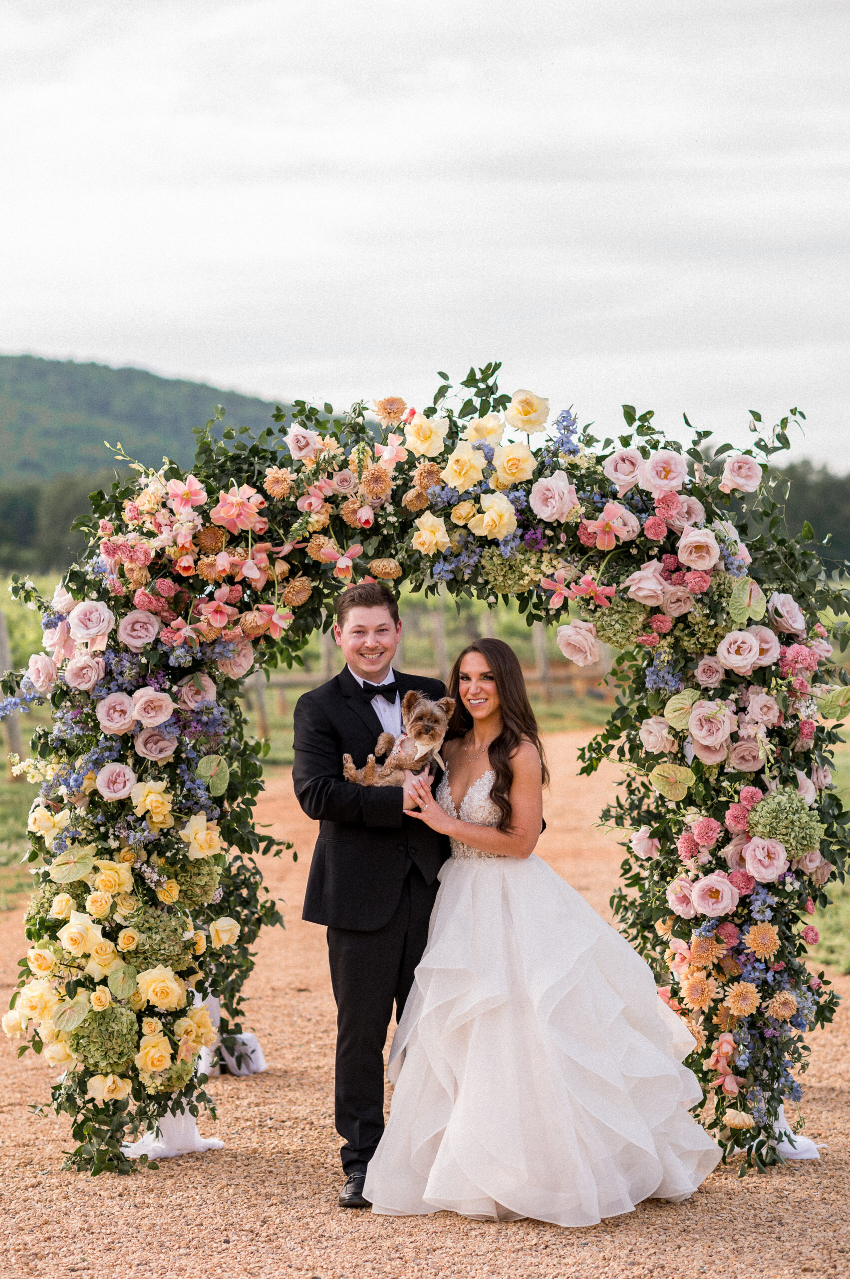 A bride and groom stand beneath a Jewish chuppah absolutely covered in beautiful florals. They're holding their adorable dog and smiling for the camera, captured beautifully by Hunter and Sarah Photography at Keswick Vineyards, on their wedding day outside Charlottesville, VA.