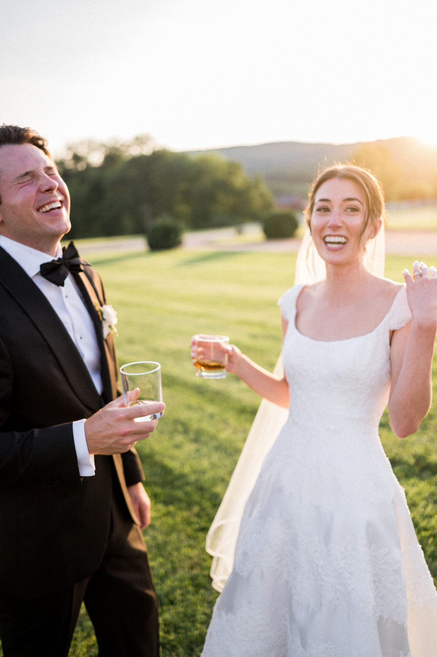 A handsome groom laughs naturally as his beautiful bride giggles, both with cocktails in their hands. The sun sets behind them, and the natural and authentic scene is beautifully captured at Castle Hill Cider by Hunter and Sarah Photography.