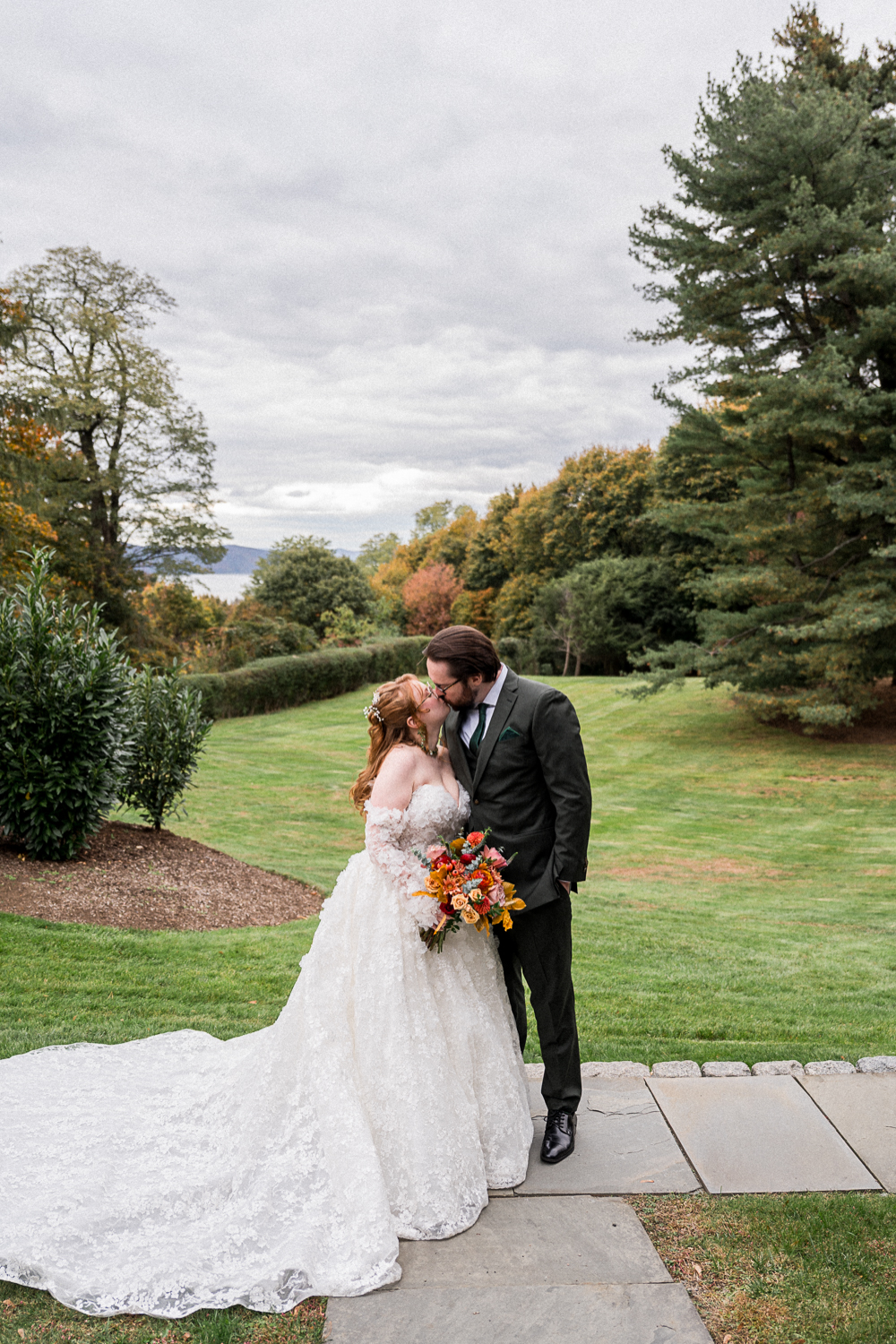 A redheaded bride and a groom in a green suit kiss with the Hudson River visible far off behind them during their Heartfelt Fall Wedding Outside New York City in Terrytown, NY. Captured by Hunter and Sarah Photography.