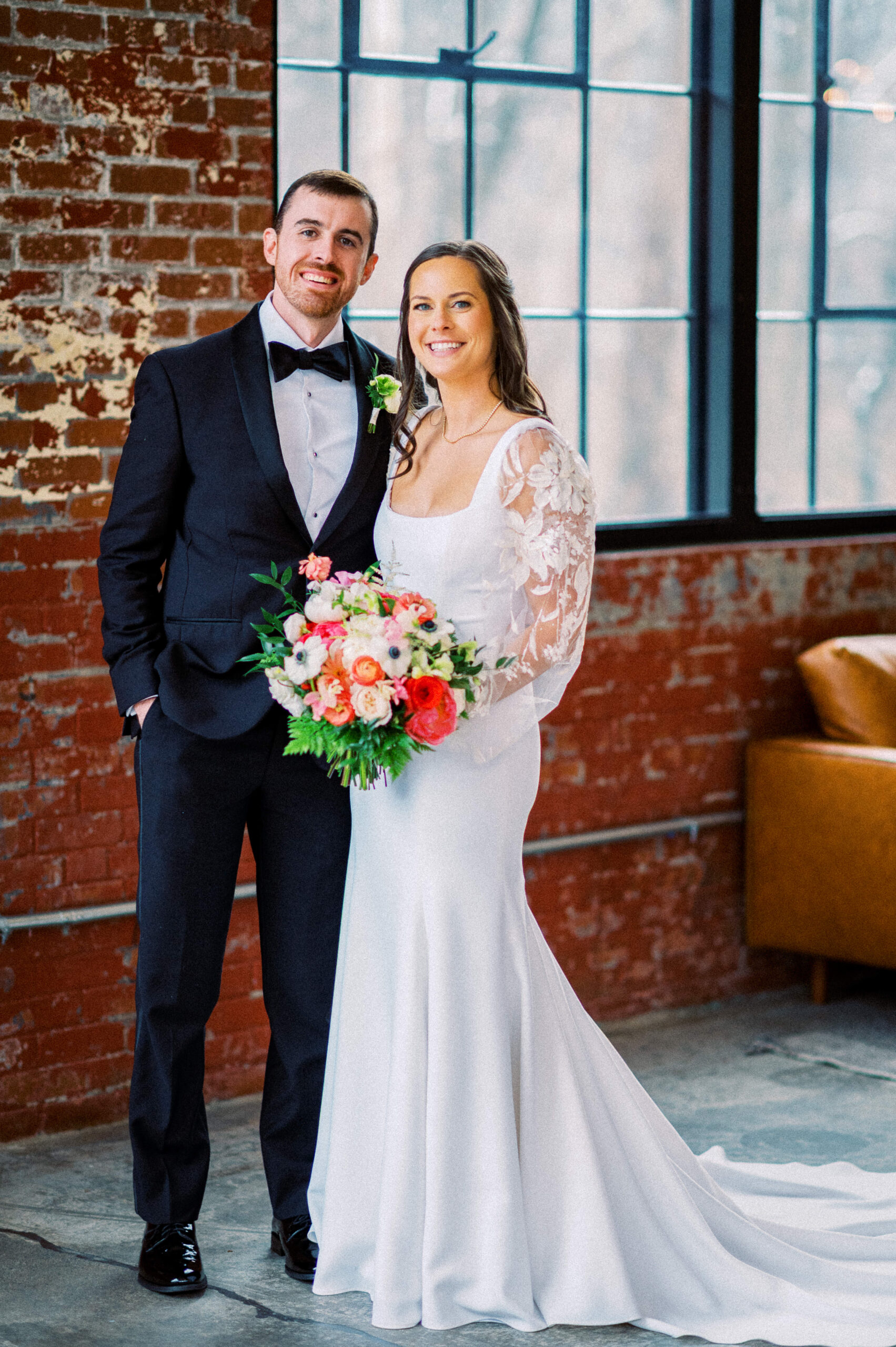 A bride and groom smile at the camera during their Wool Factory Wedding in Charlottesville, VA. Soft wintery light filters in through the windows behind them in the industrial, urban wedding venue.