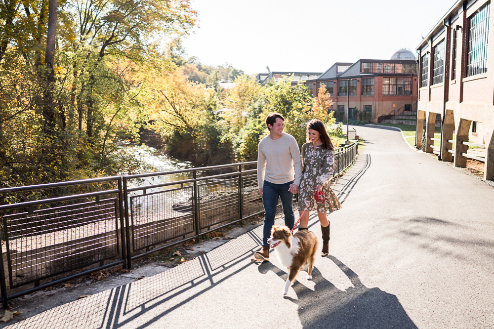 A couple walks their austrailian shepherd dog outside the Wool Factory in Charlottesville, VA. They laugh and smile during their engagement photos.