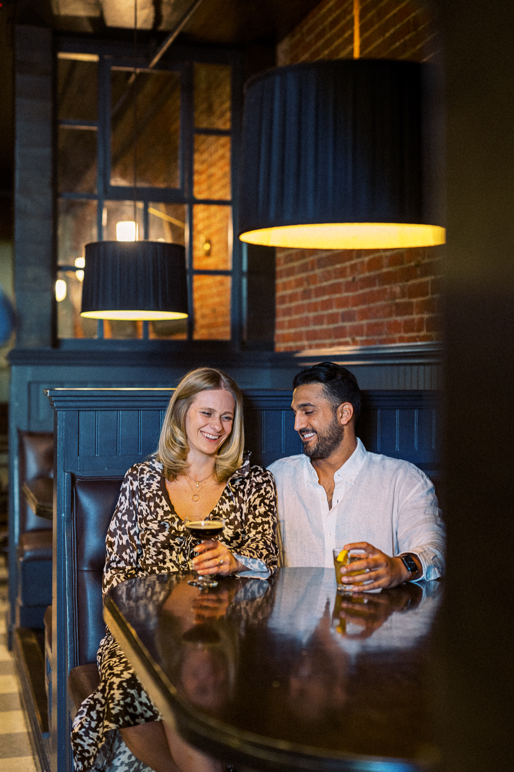 A couple laughs as they snuggle together in a booth at the Fitzroy Bar in Downtown Charlottesville, VA during their engagement photoshoot