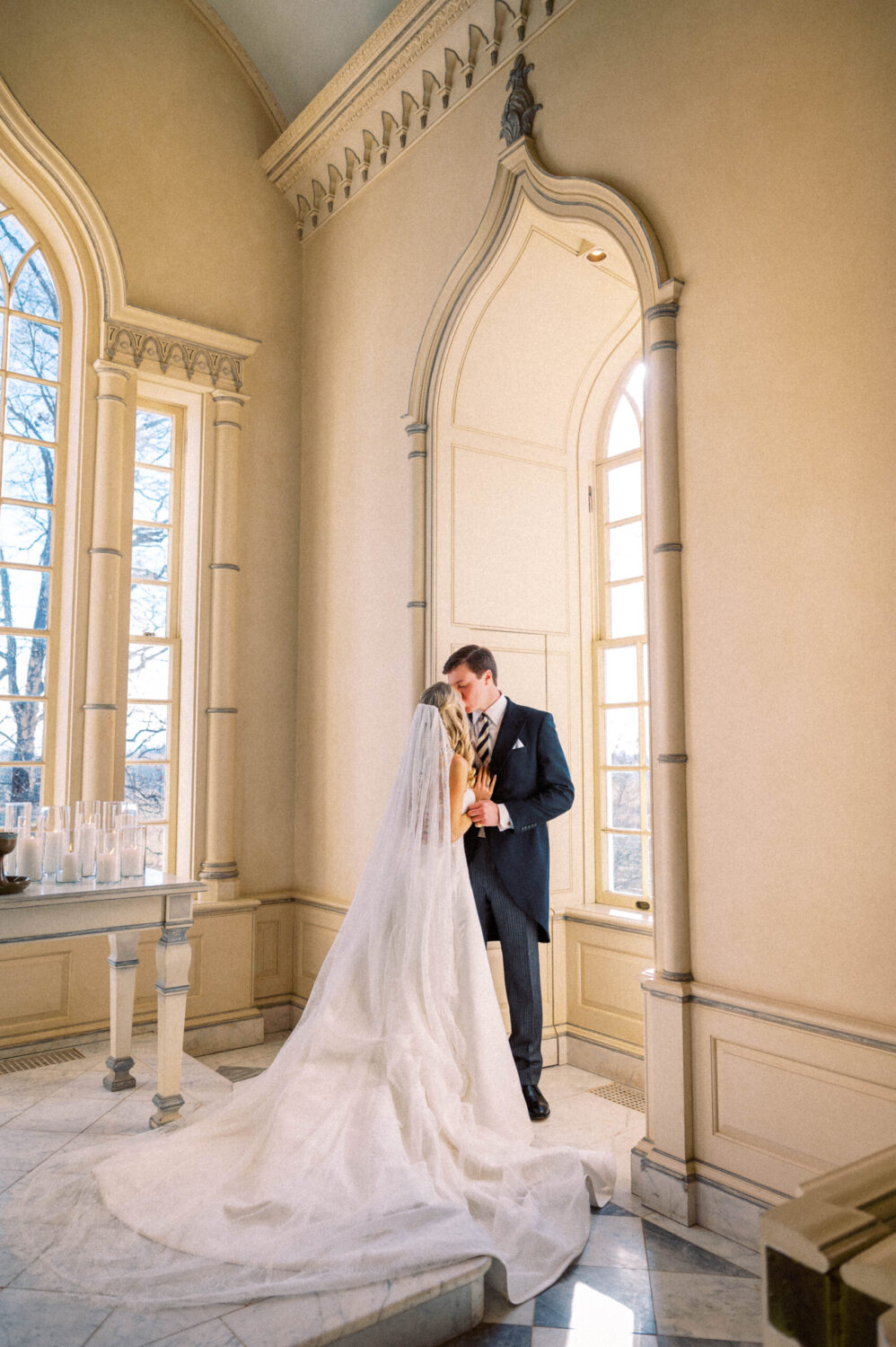 An attractive young wedding couple poses romantically inside the Trump Chapel at Albemarle Estate on their wedding day, a flowing train falls from the bride's dress and her veil. Beautifully captured by Hunter and Sarah Photography
