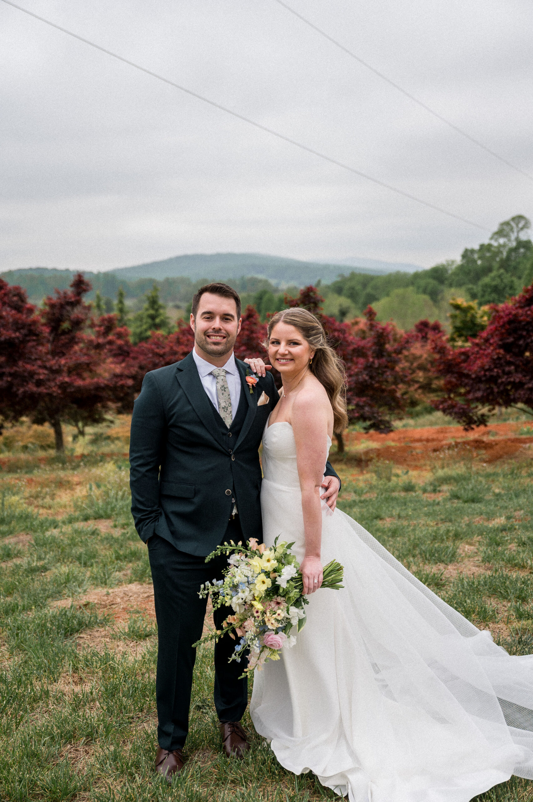 A groom in his green suit and a bride in her flowing white wedding gown and holding a gorgeous boquet of spring flowers smiles at the camera on their wedding day at the Market at Grelen. Photographed by Hunter and Sarah Photography