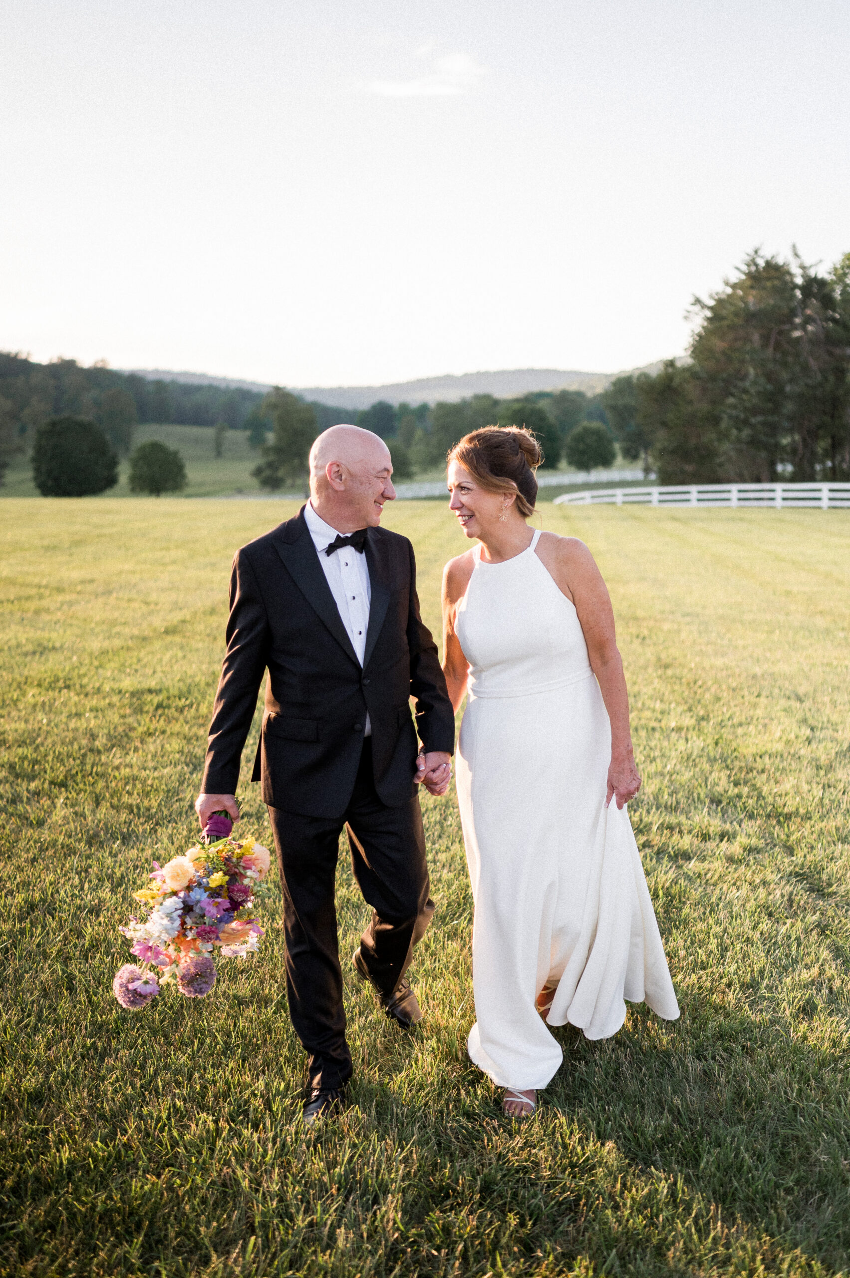 A middle-aged couple joyfully smiles at each other as they walk through a glowy field at sunset, blue ridge mountains behind them. These stunning views of a castle hill cider wedding were captured by Hunter and Sarah Photography