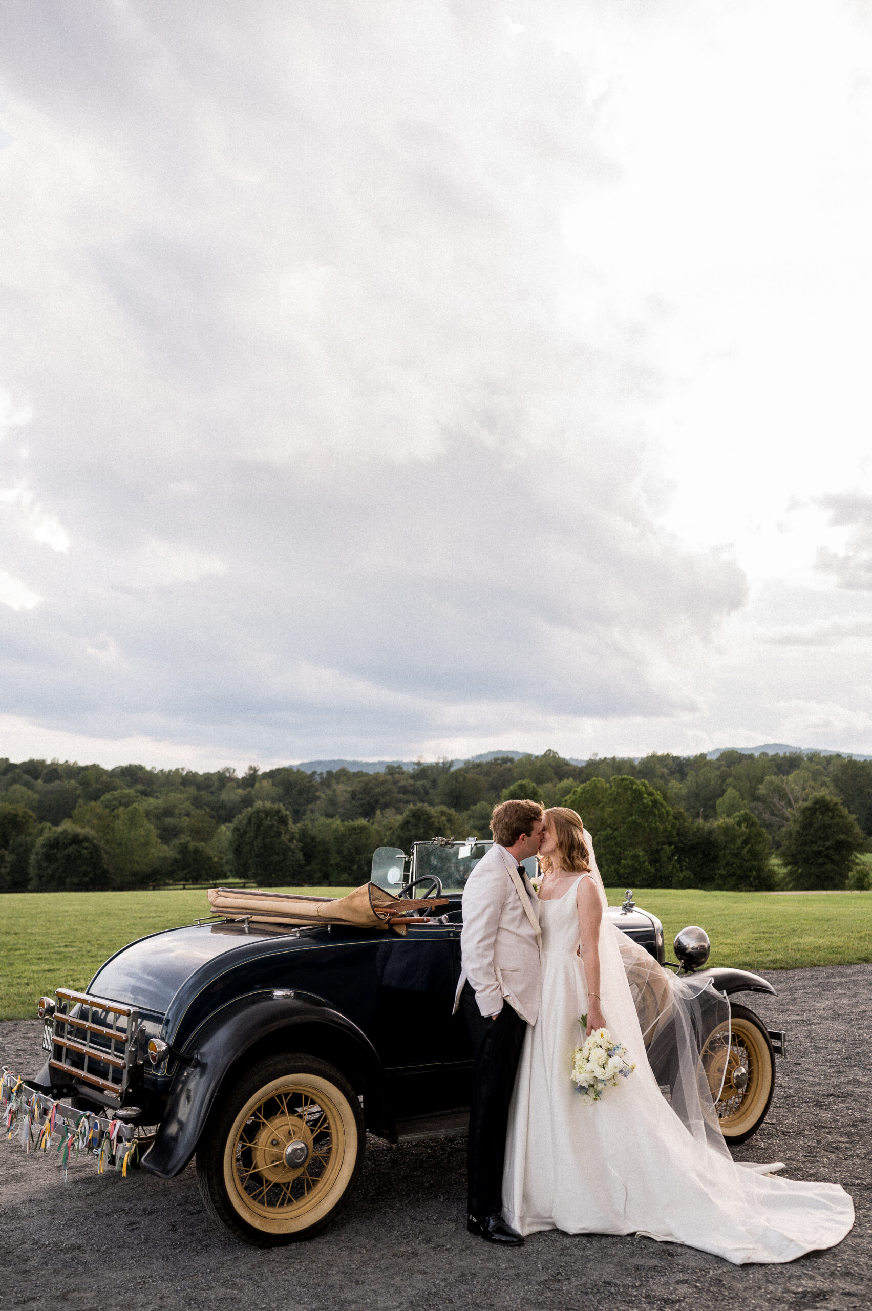 A bride in a flowing white dress kisses a groom in an ivory dinner jacket, in front of a classic Ford car during their wedding day at Tiffin Hill Farm. Captured by Hunter and Sarah Photography